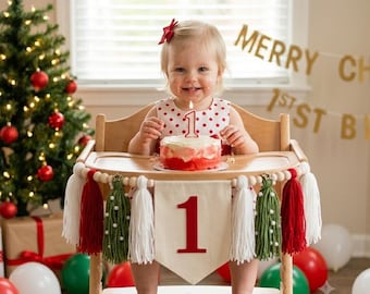 May include: A wooden high chair decorated with a banner featuring the number 1, white and red tassels, and green yarn trees. A small cake with a number 1 candle sits on the tray. The banner is made of cream fabric with a red felt number 1.