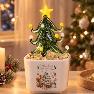May include: A glass Christmas tree ornament with green branches, yellow and white spherical accents, and a yellow star topper. It sits in a white square ceramic planter filled with pebbles, featuring a printed graphic of a squirrel, rabbit, and tree with the text "Theodore's FIRST CHRISTMAS 2026".