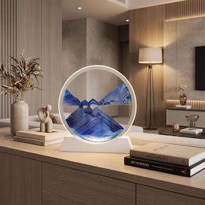 May include: A circular sand art display featuring blue and white sand layered to form mountain-like shapes within a white frame. The piece is illuminated by an integrated light ring and is supported by a solid white rectangular base.
