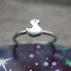 May include: A silver ring featuring a duck design. The ring is displayed against a purple and blue background.