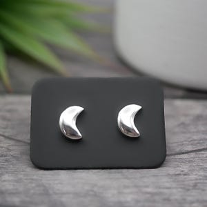 May include: Silver crescent moon stud earrings on a black display. The earrings have a polished finish, reflecting light. The background is blurred, with green foliage and a white vase. The earrings are a simple, elegant design.