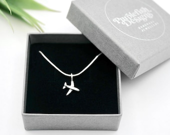 Op de afbeelding: Een zilveren vliegtuig hanger ketting gepresenteerd in een grijze geschenkdoos met het deksel open. De doos heeft een zwart interieur en de merknaam "Purplefish Designs" is op het deksel gedrukt. De ketting zit aan een witte ketting.