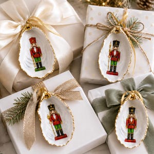 May include: Four white oyster shells, each featuring a hand-painted nutcracker figure in a red, green, and gold uniform. The shells have gold-painted edges and are attached to white gift boxes wrapped with various ribbons and decorative bows.