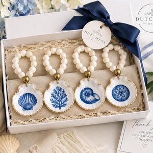 Puede incluir: Una caja de regalo blanca que contiene cuatro servilleteros, cada uno con un aro de cuentas blancas y un dije de concha de cerámica blanca con un diseño náutico azul. La caja está atada con una cinta azul marino y una etiqueta.