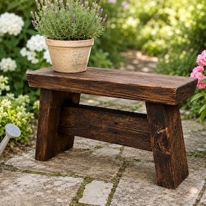 May include: A small, rectangular wooden bench with a dark brown, stained finish and visible wood grain. The bench features four sturdy, slightly angled legs connected by a horizontal support beam, providing a stable base for the flat seat surface.