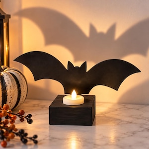 May include: A black bat-shaped silhouette mounted on a square wooden base with a small, circular, battery-operated tea light candle glowing in the center, casting a large, distinct bat-shaped shadow against the wall behind the object.