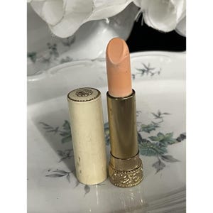 Puede incluir: Un labial con una barra de color melocotón pálido, alojado en un tubo de metal dorado con una base texturizada. La tapa cilíndrica de color blanco roto a juego presenta una parte superior dorada decorativa con un diseño floral en relieve.