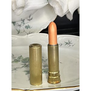 Könnte beinhalten: Ein Lippenstift mit einem schimmernden pfirsichfarbenen Stift und einer zylindrischen goldfarbenen Metallhülse mit vertikaler Riffelung und einem dekorativen gewellten Sockeldesign, abgebildet mit der passenden Kappe neben der offenen Hülse.