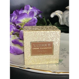 Könnte beinhalten: Eine quadratische goldfarbene Schachtel mit Ultima II Creme Foundation im Farbton Terra Rosa. Die Schachtel hat ein geprägtes Blumenmuster und ein braunes Etikett mit dem Text Ultima II, Creme Foundation, Terra Rosa und Net Wt 0.8 oz.