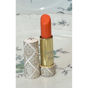 Puede incluir: Un tubo de pintalabios naranja brillante con una carcasa de metal dorado. La tapa extraíble y la base del tubo presentan un patrón de damasco blanco y tostado. El texto Moon Drops Lipstick Revlon está impreso en el tubo de metal dorado.