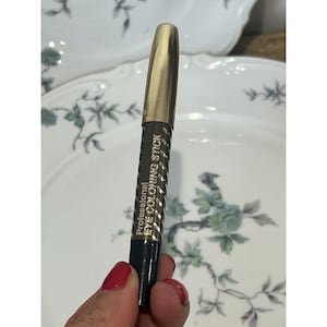 May include: A cylindrical eye colouring stick with a metallic gold cap and a black body featuring diagonal silver stripes. The text "Professional EYE COLORING STICK" is printed in white on the side of the black barrel of the cosmetic applicator.