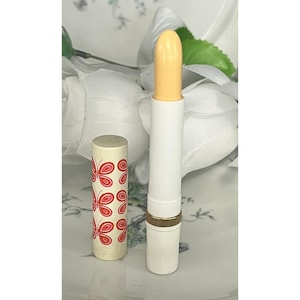 Op de afbeelding: Een cilindervormige cosmetische stick met een lichtgele balsem en een witte basis. De afneembare dop is wit en voorzien van een herhalend patroon van kleine rode vlinders. De huls heeft een dunne goudkleurige ring tussen de basis en de bovenkant.