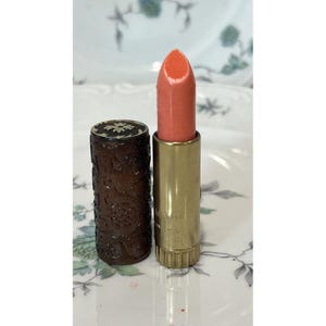 Peut inclure: Un rouge à lèvres avec un bâton couleur pêche et une base en métal doré. Le capuchon cylindrique assorti est marron foncé avec un motif floral en relief et un petit motif en forme d'étoile sur le dessus. La base présente un bord inférieur cannelé.