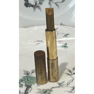 Op de afbeelding: Een vintage goudkleurige metalen lippenstiftkoker met een cilindrische vorm en een verticaal geribbelde textuur. De koker is geopend met de lippenstift uitgedraaid en de bijbehorende losse dop staat rechtop naast de basis van de koker.