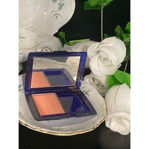 May include: A rectangular navy blue makeup compact case with an open lid revealing a mirror and a single square pan of peach-colored powder blush. The compact features a hinged design and a smooth, glossy finish on the exterior casing.