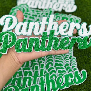 Pode incluir: Uma pilha de emblemas bordados com a palavra Panthers em fonte cursiva. O emblema superior tem letras brancas com borda verde, enquanto o emblema abaixo apresenta letras verdes com borda branca, ambos com um acabamento texturizado.