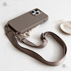 May include: A taupe-colored smartphone case with a matching adjustable nylon crossbody strap. The case features a cutout for a triple-lens camera system and side buttons. The strap attaches to the case with two metal clips and includes a plastic slider for length adjustment.