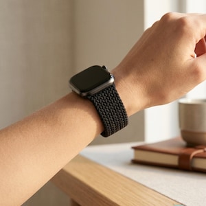 May include: A black smartwatch featuring a rectangular screen and a textured, braided elastic watch band. The band has a woven, herringbone-like pattern that wraps around the wrist, providing a flexible and secure fit for the electronic device.