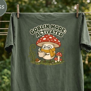 May include: A moss green t-shirt featuring a graphic of a cartoon mushroom character wearing glasses and a yellow scarf, holding a steaming mug. The text above reads GOBLIN MODE: ACTIVATED. A small snail and two smaller mushrooms are also depicted.