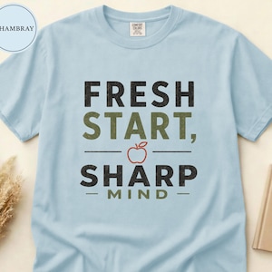 May include: A light blue short-sleeve t-shirt featuring the text "FRESH START, SHARP MIND" in black and olive green lettering. A small red apple graphic is centered between the two lines of text. The shirt has a crew neckline and a tag.