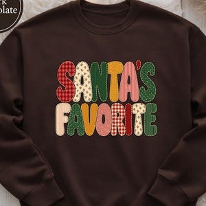 May include: A dark chocolate brown crewneck sweatshirt featuring the text SANTA'S FAVORITE in a bold, rounded font. Each letter is filled with a different festive pattern, including red and white gingham, green polka dots, and gold star prints.