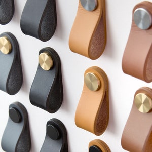 Peut inclure: Assortiment de poignées de tiroir en cuir avec des accents en laiton. Les poignées sont de différentes couleurs, notamment noir, marron et beige.