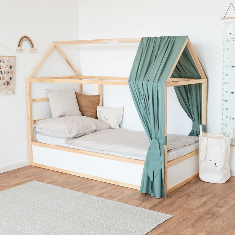 Ein Ikea Kura Kinderbett wurde am Fußende mit einem grünen Vorhang aus Baumwolle dekoriert. Im Bett liegen Kissen und andere Textilien.