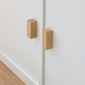 Può includere: Due pomelli per armadio in legno, di forma rettangolare, sono montati su una porta di armadio bianca. I pomelli sono in legno marrone chiaro e hanno una finitura liscia.