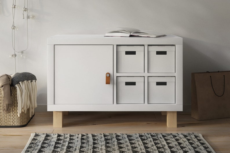 Ein 2er Ikea Kallax Regal in weiß steht auf eckigen Möbelfüßen aus Buchenholz. Das Regal ist mit einer Tür und einem Regalkreuz gefüllt, in dem 4 Schubladen stecken.