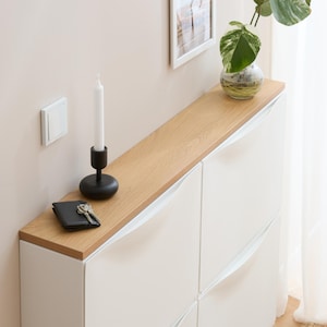 May include: A white cabinet with a light wood top. A black candle holder with a white candle, a black wallet, and keys sit on the top. A small glass vase with a plant is also on the top. A framed picture hangs on the wall.