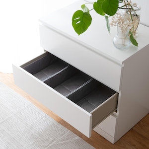 Op de afbeelding: Witte commode met drie lades, de bovenste lade is open en heeft grijze stoffen dividers erin.