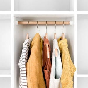 Könnte beinhalten: Fünf verschiedene Pullover hängen an Holzbügeln in einem weißen Schrank. Die Pullover sind in verschiedenen Farben, darunter Gelb, Orange, Braun und Weiß.