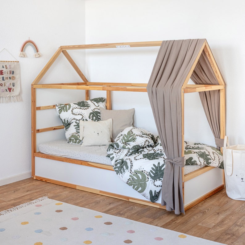 Ein Ikea Kura Hausbett mit Dach wurde mit einem Baldachin aus Musselinstoff in der Farbe beige dekoriert. Zudem liegen im Bett Kissen und Bettdecke.