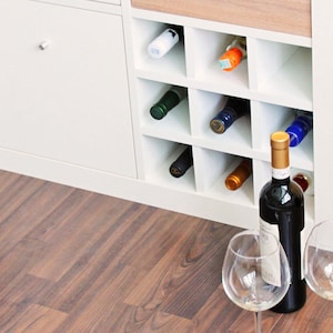 Weißer Ikea Kallax Expedit Regaleinsatz für 9 Flaschen für ein Ikea Kallax Regal. In 8 Fächern stecken Weinflaschen. Vor dem Regal steht eine Weinflasche mit 2 Weingläsern.
