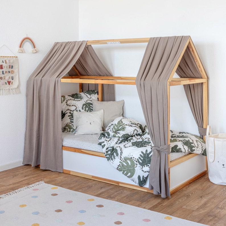 Ein Ikea Kura Hausbett mit Dach wurde mit zwei Baldachinen aus Musselinstoff in der Farbe beige dekoriert. Zudem liegen im Bett Kissen und Bettdecke.