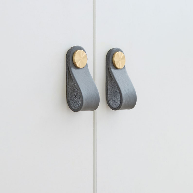 2 stylische Möbelgriffe aus Leder in Anthrazit mit je 2 goldenen Metallknöpfen, die an weißen Türen befestigt sind.