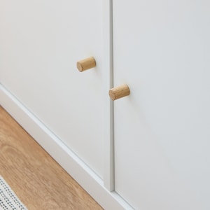 Könnte beinhalten: Nahaufnahme eines weißen Schranks mit zwei zylindrischen Holzknöpfen. Die Knöpfe sind aus hellfarbigem Holz gefertigt und an den Schranktüren angebracht. Der Schrank steht auf einem hellfarbigen Holzboden.