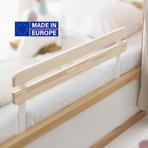 Op de afbeelding: Houten bedhek met een lichte houtafwerking en witte steunen. Bevestigd aan een bedframe met een witte matras en beddengoed. De afbeelding bevat de tekst "MADE IN EUROPE" met de vlag van de Europese Unie.