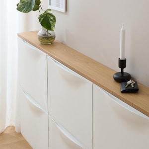 Könnte beinhalten: Ein weißer Schrank mit einer hellen Holzplatte. Links steht eine kleine Glasvase mit einer grünen Pflanze. Rechts befinden sich ein schwarzer Kerzenhalter mit einer weißen Kerze und ein schwarzes Portemonnaie mit Schlüsseln.