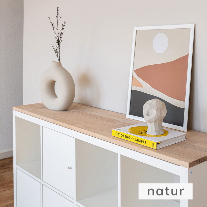 Eine Holzplatte aus naturbelassenem Buchenholz liegt auf einem weißen 8er Ikea Kallax Regal. Auf der Holzplatte stehen Deko-Artikel, wie ein Bild, eine Vase und ein Buch.