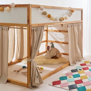 Op de afbeelding: Een houten hoogslaper met beige gordijnen en een kleurrijk geometrisch tapijt. Het bed is versierd met lichtslingers en een slinger van ronde stoffen lampjes. Een pluche kikkerkussen en een houten speelgoedtreinset liggen in het bed.