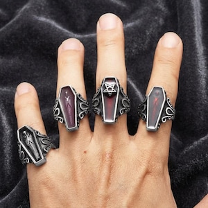 May include: Four silver-toned metal rings shaped like coffins, each featuring intricate scrollwork on the bands. Three rings contain dark red enamel inlays, with one displaying a silver pentagram symbol, while the fourth ring features a silver skeleton figure.