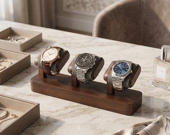 May include: A dark wood watch stand featuring three individual cushioned mounts for displaying wristwatches. The stand holds three distinct watches: one with a brown leather strap, one with a silver metal link band, and one with a silver metal band.