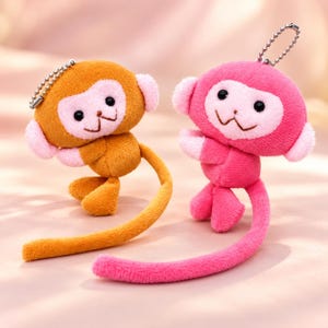 May include: Two plush monkey keychains, one in golden brown and one in bright pink. Each monkey features a light pink face, small round ears, black bead eyes, a stitched mouth, and a long, slender tail. A silver metal ball chain is attached to the top.