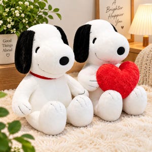 May include: Two white plush dog toys with black ears and noses. One toy wears a red collar, while the other holds a large, textured red heart. Both have stitched eyes and mouths, sitting upright on a soft, cream-colored surface with visible seams.