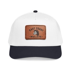Puede incluir: Una gorra de béisbol blanca con visera azul marino oscuro y un botón superior. En el centro de la parte frontal hay un parche rectangular de cuero marrón con un gráfico negro de un pez con corte mullet y el texto EAST COAST MULLETS.