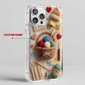 May include: A clear phone case featuring a printed design of a brown woven basket filled with colorful yarn balls, knitting needles, and a crochet hook. The design includes a small beige hat, red and beige hearts, stitch markers, and the name Ann.