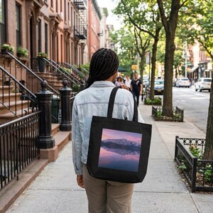 May include: A black tote bag featuring a square graphic print of a mountain range reflected in a calm lake at sunset with pink and purple hues. The bag has two shoulder straps and is worn over the shoulder with a light blue denim jacket and pants.