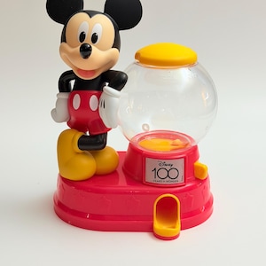 May include: A Mickey Mouse gumball machine featuring a red base, a clear spherical container with a yellow lid, and a white, black, red, and yellow figure of Mickey Mouse. The front displays a Disney 100 Years of Wonder logo on a small plaque.
