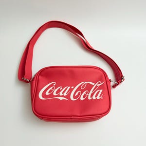 Könnte beinhalten: Eine leuchtend rote, rechteckige Umhängetasche mit dem weißen Coca-Cola-Logo auf der Vorderseite. Die Tasche verfügt über einen passenden, verstellbaren roten Schulterriemen mit silberfarbenen Metallbeschlägen und einen Reißverschluss.